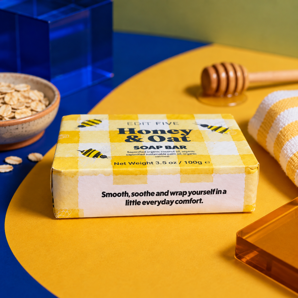 A bar of Edit Five Honey &amp; Oat soap bar in a yellow and white gingham box, styled on a yellow and blue surface with honey-themed props including a wooden dipper, honeycomb, oats, and a folded yellow-and-white cloth. Edit Five honey and oat soap bar featuring nourishing honey and comforting oats, beautifully packaged as a thoughtful pampering gift, bathroom essential or gift-hamper addition.