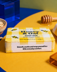 A bar of Edit Five Honey & Oat soap bar in a yellow and white gingham box, styled on a yellow and blue surface with honey-themed props including a wooden dipper, honeycomb, oats, and a folded yellow-and-white cloth. Edit Five honey and oat soap bar featuring nourishing honey and comforting oats, beautifully packaged as a thoughtful pampering gift, bathroom essential or gift-hamper addition.