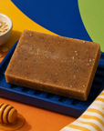 A rectangular bar of oatmeal shampoo or soap in a medium brown color with visible oat flakes, resting on a blue tray. The scene is styled with a small bowl of oats, a wooden honey dipper with honey, and a striped cloth, set against a colorful background of yellow, blue, and green shapes. Handmade 100g artisan honey and oatmeal soap, created for a mild, soothing wash with a creamy lather—ideal for daily bathing, handwashing and simple self-care routines.