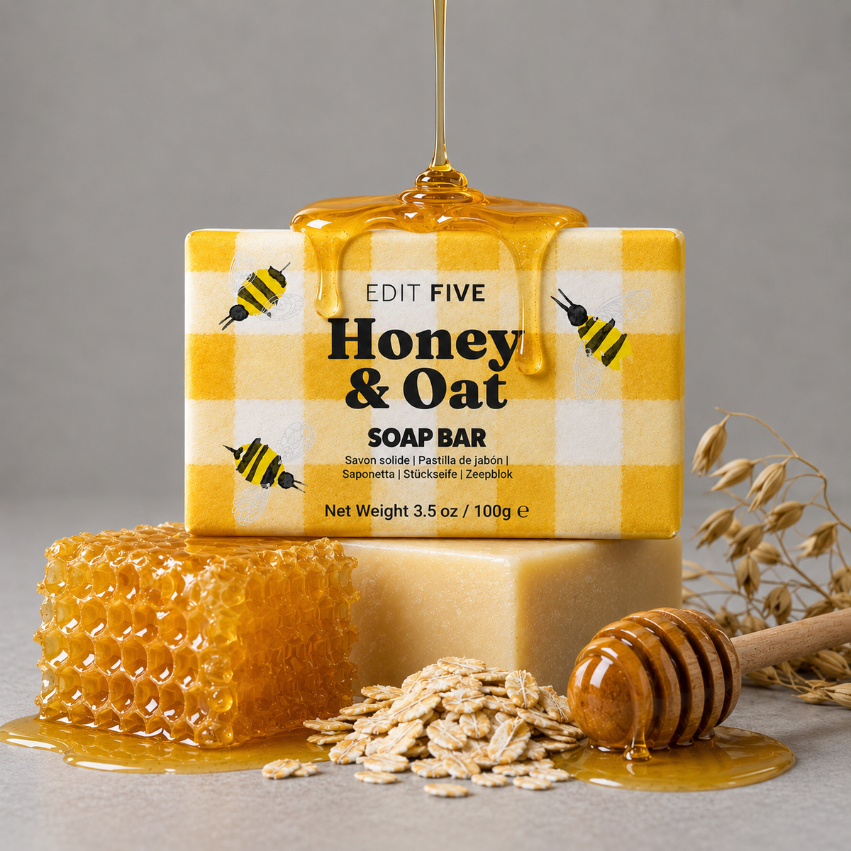 A boxed Honey &amp; Oat soap bar sits on top of a plain soap bar, with honey dripping from above. The front box is yellow-and-white checkered with bumblebee illustrations and the text 'EDIT FIVE Honey &amp; Oat SOAP BAR. Luxury handmade honey and oatmeal soap with a gentle cleansing formula, soft natural fragrance and creamy lather, perfect for anyone who enjoys comforting bath and body products.