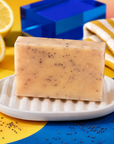 A rectangular bar of pale beige soap with small dark specks sits on a white ribbed dish. The scene is styled with lemon slices, a blue box, and a yellow-and-white striped cloth on a bright yellow and blue surface. Handmade 100g artisan lemon soap combining uplifting citrus essential oil with natural poppy seeds for a fresh fragrance, creamy lather and gentle everyday exfoliation.