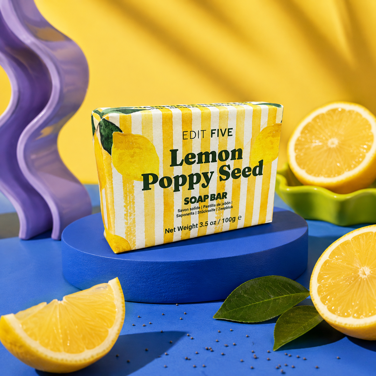 A bar of Edit Five Lemon Poppy Seed soap is displayed on a blue pedestal, surrounded by fresh lemon slices, green leaves, and a purple decorative prop against a bright yellow background. Luxury handmade citrus soap with refreshing lemon essential oil and gently exfoliating poppy seeds, designed to cleanse, buff and revitalise the skin during everyday use.