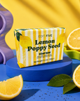 A bar of Edit Five Lemon Poppy Seed soap is displayed on a blue pedestal, surrounded by fresh lemon slices, green leaves, and a purple decorative prop against a bright yellow background. Luxury handmade citrus soap with refreshing lemon essential oil and gently exfoliating poppy seeds, designed to cleanse, buff and revitalise the skin during everyday use.