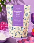 A stand-up pouch of Edit Five Moonlight Soak bath salts, featuring a lavender label and a cream background with blue jellyfish and starfish illustrations. The pouch is styled on a purple surface with a pink glass dish of white bath salts, folded pastel towels, and a purple candle in the background. Vegan-friendly lavender bath salts handmade in Staffordshire with mineral-rich Dead Sea salts, Epsom salts and uplifting bergamot and sweet orange essential oils.