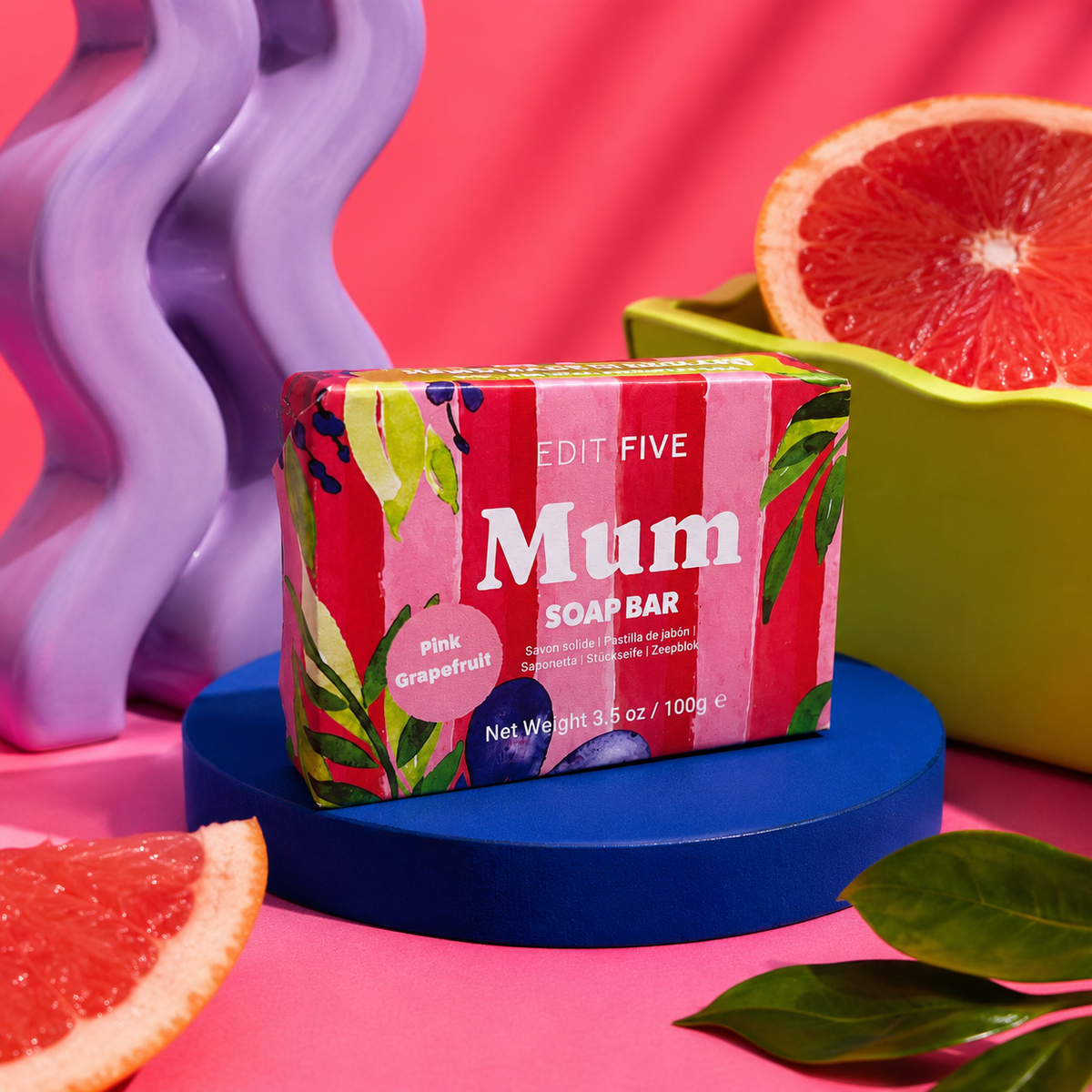 A pink grapefruit scented soap bar by Edit Five, packaged in a colorful box with botanical illustrations, displayed on a blue pedestal with grapefruit slices and a purple sculptural background. Luxury handmade soap for Mum with refreshing pink grapefruit essential oil, a rich everyday lather and colourful blush packaging, perfect for Mother’s Day and birthday gifting.
