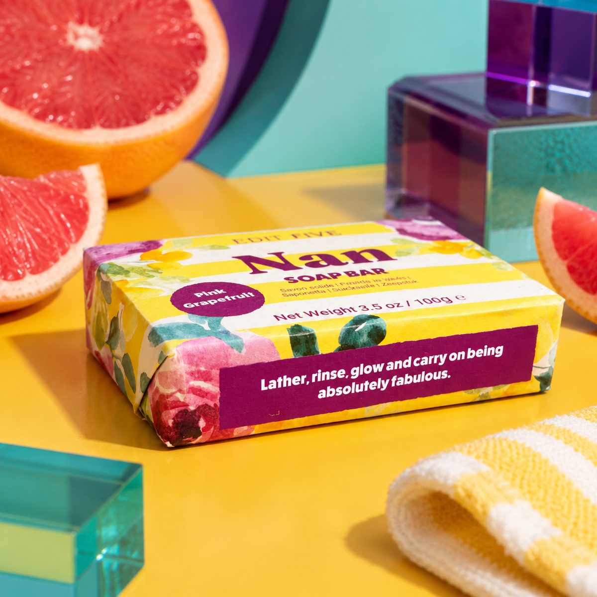 A wrapped bar of soap bar sits on a bright yellow surface with grapefruit slices, teal blocks, and a folded yellow-and-white towel around it. The soap packaging is floral and reads 'Nan SOAP BAR' and 'pink grapefruit. Edit Five pink grapefruit soap bar designed especially for Nan, featuring natural essential oils and beautiful packaging—ideal as a pampering gift, self-care present or gift-box addition.
