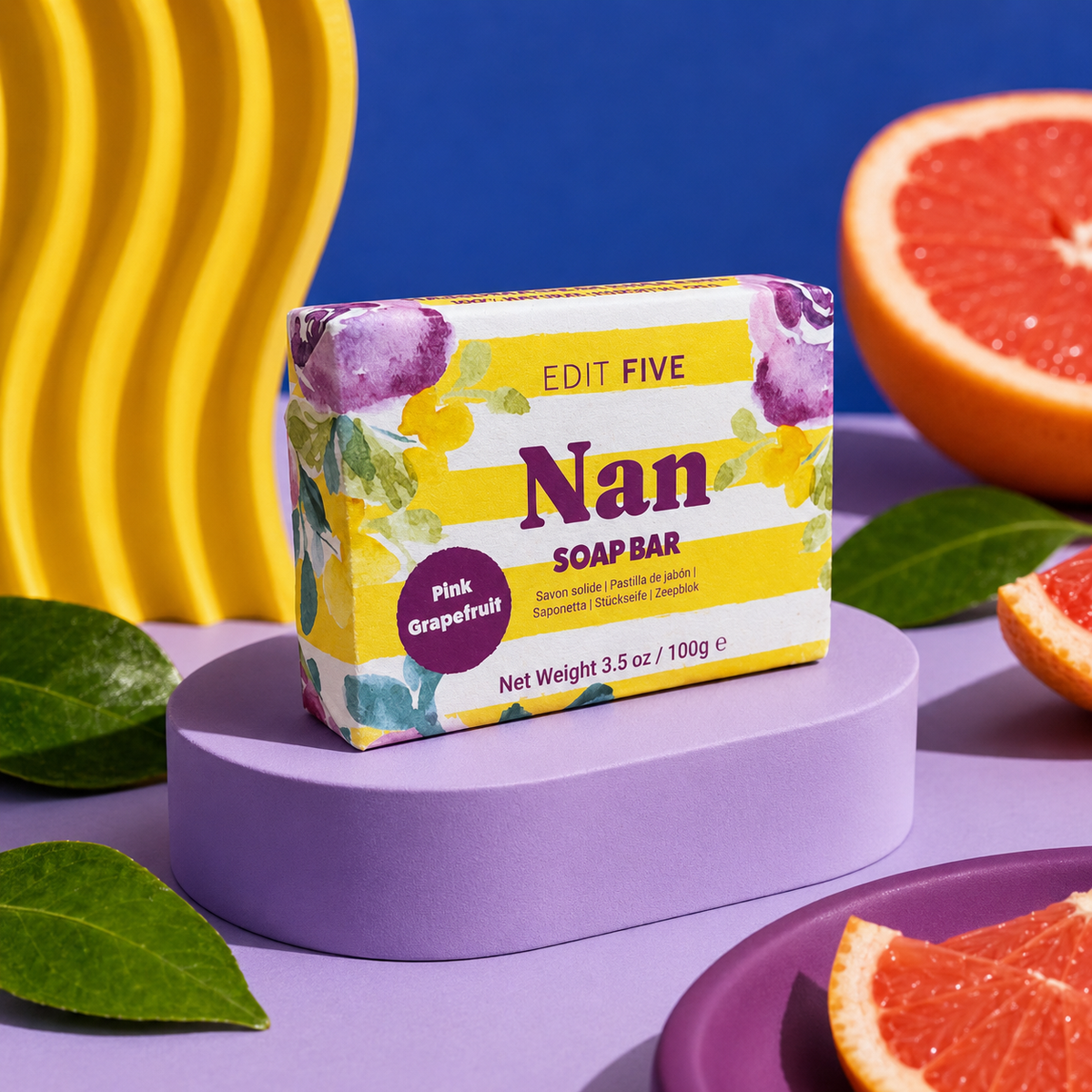 A bar of soap from the brand Edit Five, specifically a Pink Grapefruit scent, displayed on a purple pedestal. The packaging is yellow and white with purple and green floral graphics. Nan pink grapefruit soap bar, handmade in England with 100% natural essential oil and colourful gift-ready packaging—a thoughtful birthday, Christmas or Mother’s Day gift for Nan.