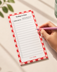 A hand with pink nails holds a lavender pen over a red-and-white checkered notepad labeled 'Today's List' on a light beige surface with soft shadows and a green glass in the background. Colourful “Today’s List” long daily planner notepad with lined sections for organising tasks, priorities and reminders. A bright, modern desk stationery essential for productivity, work, home organisation, study and everyday to-do lists.