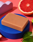 A rectangular bar of light tan soap sits on a round blue pedestal, surrounded by grapefruit slices, green leaves, a small green dish, and a lavender sculptural object on a pink background. Handmade 100g artisan soap for Mum, scented with uplifting pink grapefruit essential oil for a bright, fruity fragrance and refreshing everyday cleansing experience.