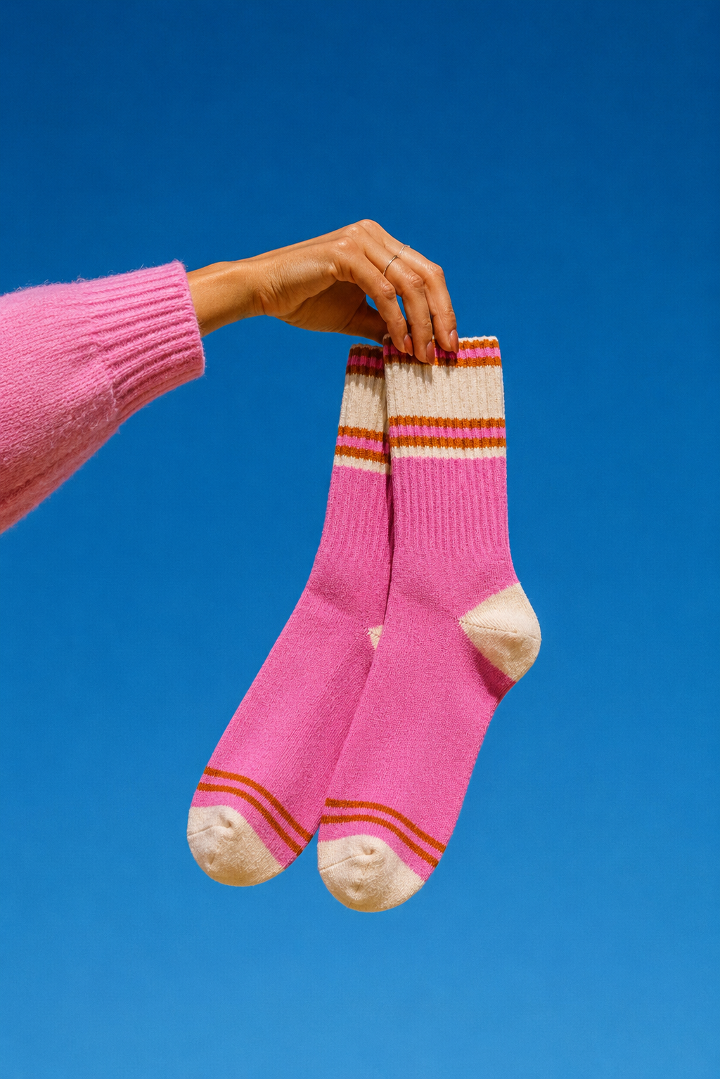A person's hand wearing a pink knit sweater holds up a pair of pink knit socks with cream toe, heel, and cuff details, set against a solid blue sky. Colourful women’s wool-blend socks featuring vibrant pink, peach and orange stripes—a fun fashion accessory, cosy winter essential and thoughtful gift for her.