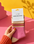 A hand wearing a rust-colored knit sweater holds a pair of pink and beige striped socks with a white paper tag that reads 'EDIT FIVE SIGNATURE SOCKS'. The background is a vibrant pink and yellow studio setup with leaf shadows. Women’s pink, peach and orange striped socks made from a soft, warm wool-blend fabric, with a comfortable stretch fit for colourful everyday wear.