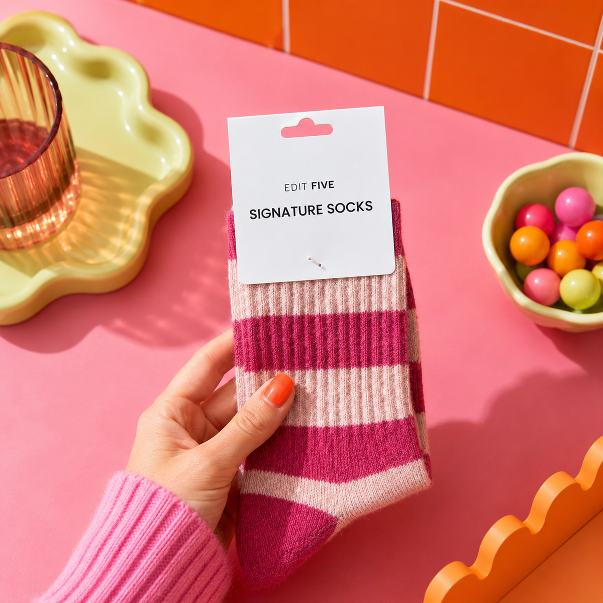 A hand holds a pair of pink and cream striped socks with a white paper tag that reads 'EDIT FIVE SIGNATURE SOCKS' against a bright pink background with colorful props. Women’s cherry pink and baby pink striped socks made from a soft, warm and breathable wool-blend fabric, with a comfortable stretch fit for cosy everyday wear.