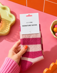 A hand holds a pair of pink and cream striped socks with a white paper tag that reads 'EDIT FIVE SIGNATURE SOCKS' against a bright pink background with colorful props. Women’s cherry pink and baby pink striped socks made from a soft, warm and breathable wool-blend fabric, with a comfortable stretch fit for cosy everyday wear.