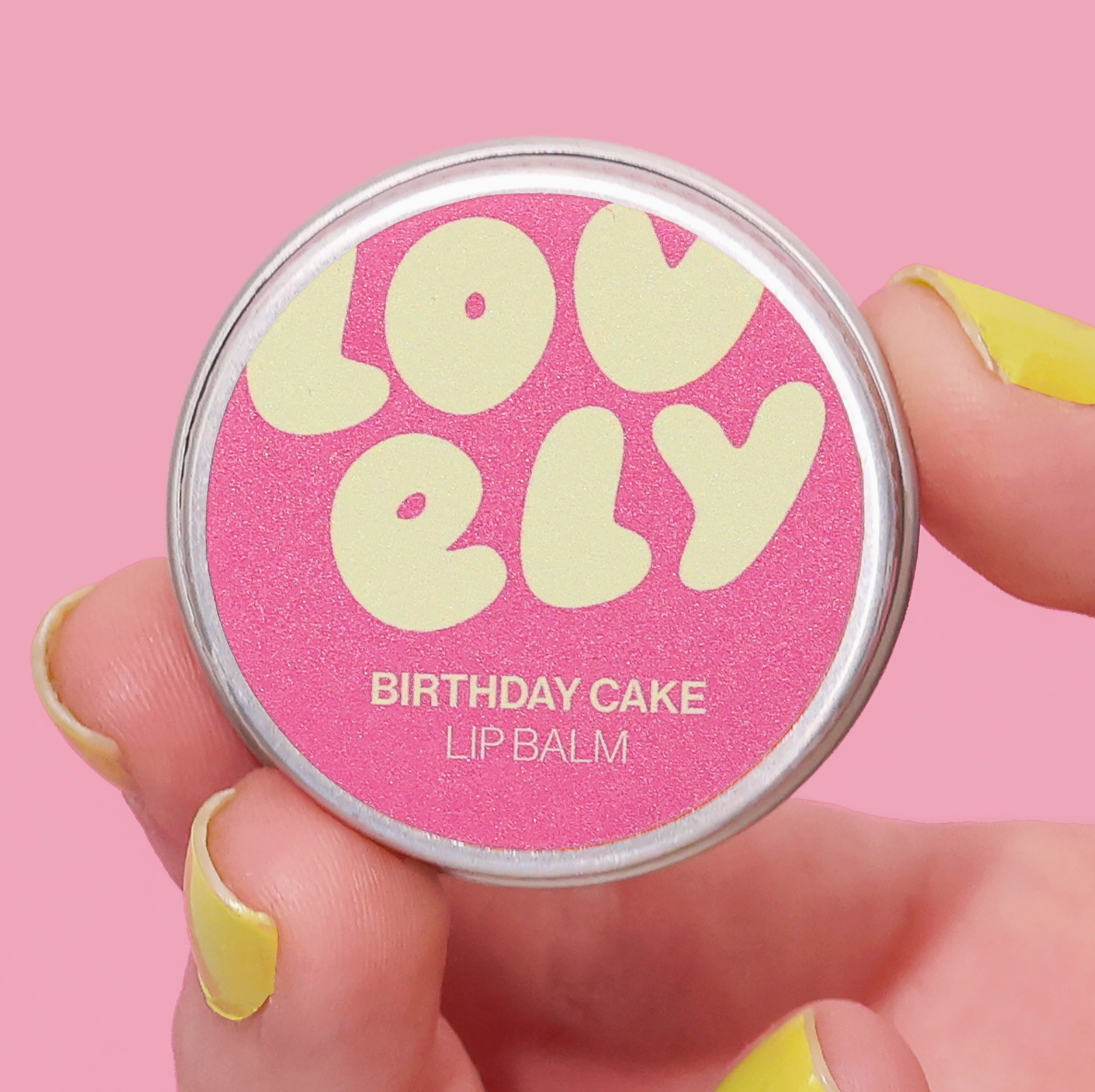 Lip balm container labeled 'Lovely Birthday Cake' held by a hand with yellow nail polish against a pink background.