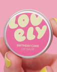 Lip balm container labeled 'Lovely Birthday Cake' held by a hand with yellow nail polish against a pink background.