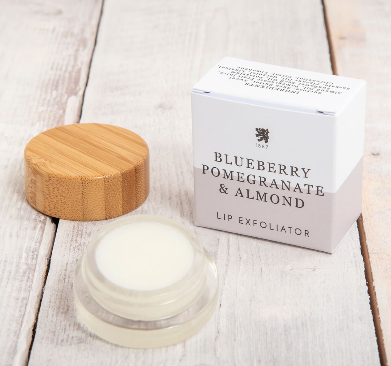 Lip exfoliator with wooden cap and packaging labeled 'Blueberry Pomegranate & Almond' on a light wooden surface.