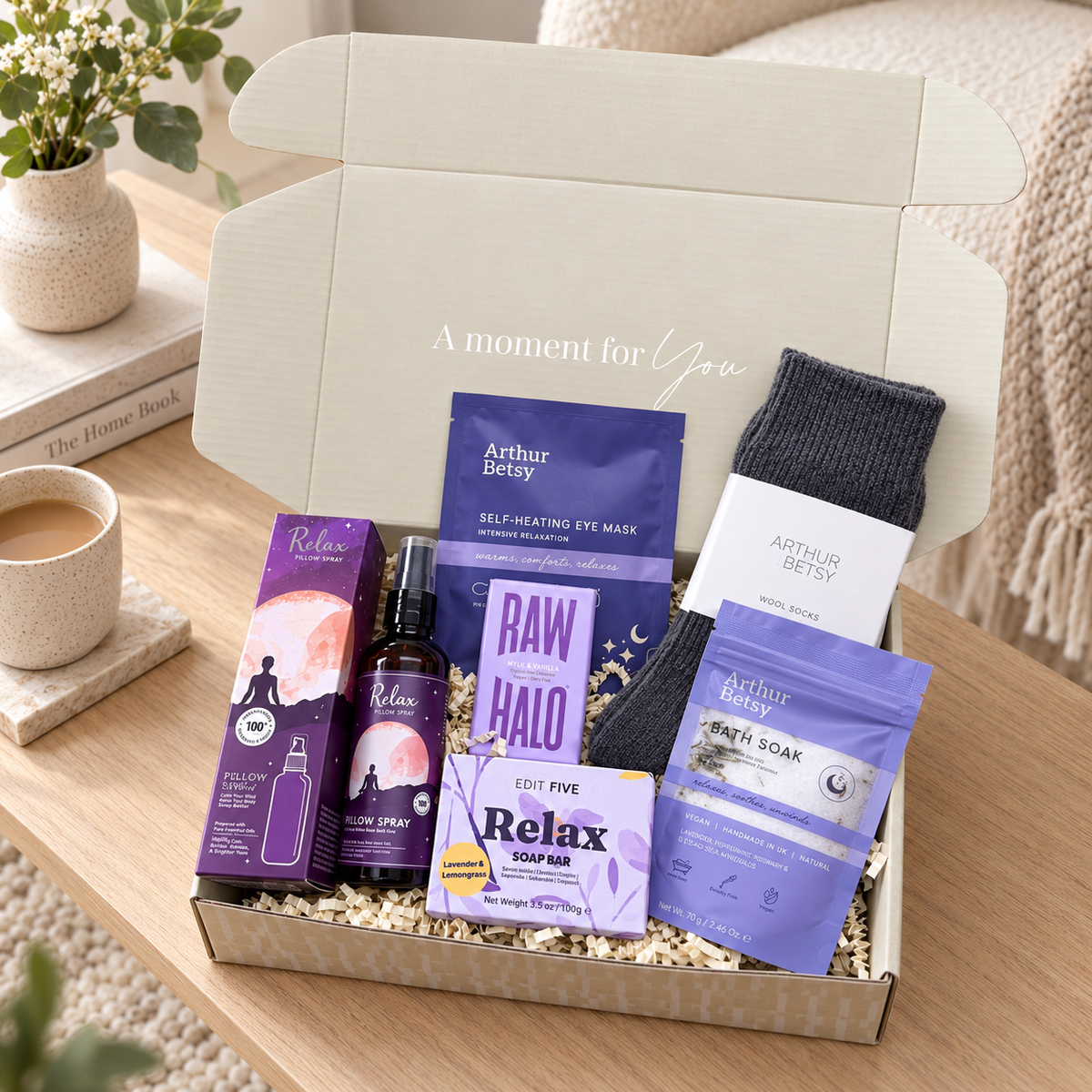 A curated self-care gift box set on a wooden table, featuring Arthur Betsy products including a self-healing eye mask, eye cream, bath soak, and soap, alongside a dark gray knit sock, all arranged in a kraft box with shredded paper filler.