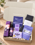 A curated self-care gift box set on a wooden table, featuring Arthur Betsy products including a self-healing eye mask, eye cream, bath soak, and soap, alongside a dark gray knit sock, all arranged in a kraft box with shredded paper filler.
