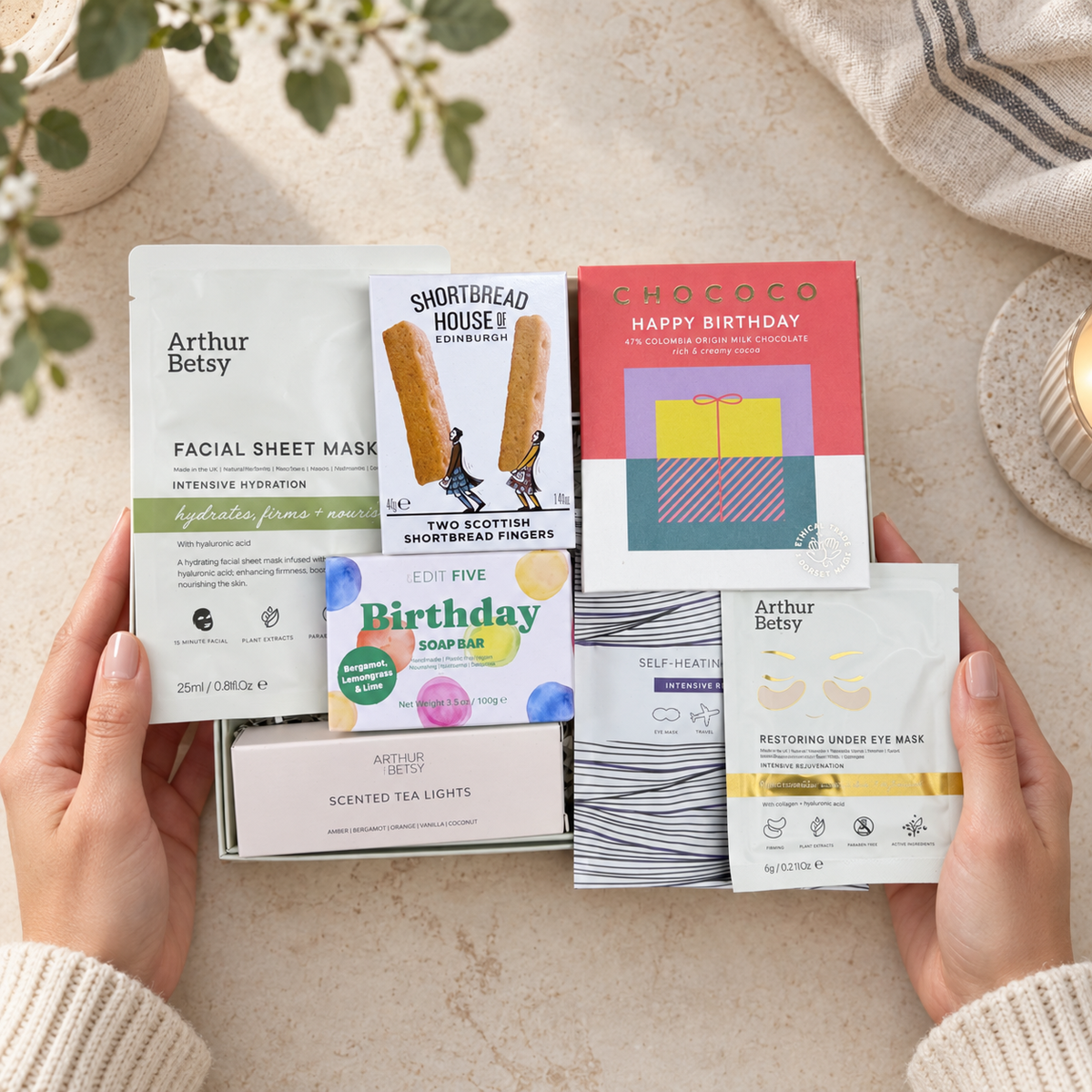 A person's hands hold a curated gift set of skincare and tea accessories against a warm beige background with soft plant and fabric props. The set includes Arthur Betay facial sheet masks, a boxed soap bar, a colorful 'Happy Birthday' card, a notebook, and scented tea lights. Hallby birthday letterbox gift for her with soap, face masks, scented tealights, warming eye mask and chocolate