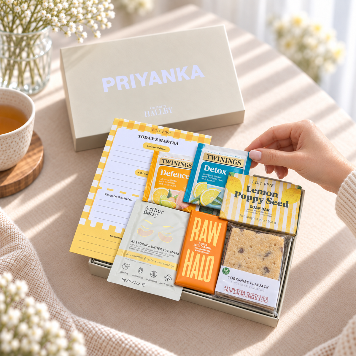 A gift box arrangement on a beige tablecloth with a hand placing a yellow card into a silver tray. The box contains tea packets, a notepad, a card, and a cookie, with a large beige gift box labeled PRIYANKA behind it. Pick me up gift for her featuring Today’s Mantra notepad, sunshine coaster and relaxing self-care treats
