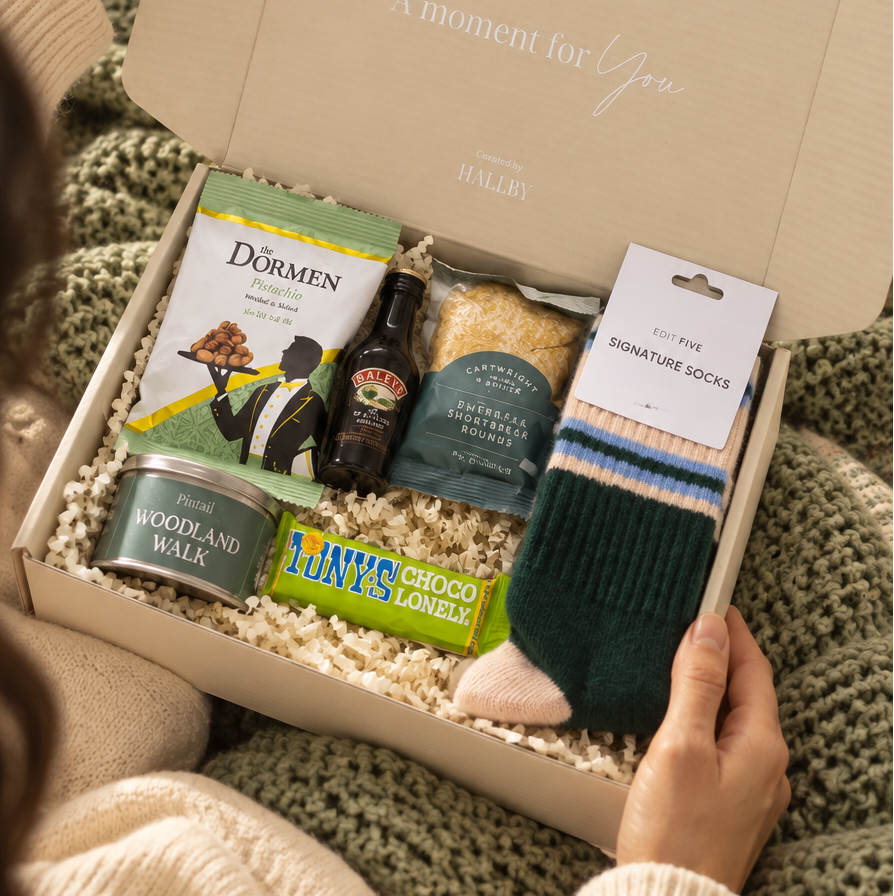 A cozy gift box filled with assorted treats and socks, held open by a person wearing a cream sweater over a green knit blanket. Visible items include Dorchen cookies, a bottle of dark rum, a can of stout, a bag of Lay's chips, and a pair of striped socks. Hallby Christmas gift box for men with Edit Five socks, Woodland Walk candle tin, Baileys and festive snacks
