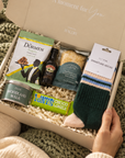 A cozy gift box filled with assorted treats and socks, held open by a person wearing a cream sweater over a green knit blanket. Visible items include Dorchen cookies, a bottle of dark rum, a can of stout, a bag of Lay's chips, and a pair of striped socks. Hallby Christmas gift box for men with Edit Five socks, Woodland Walk candle tin, Baileys and festive snacks
