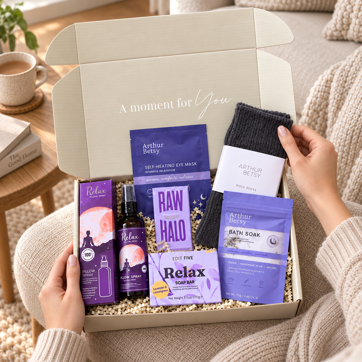 A curated self-care gift box opened on a cozy sofa, containing purple and lavender skincare items, a dark gray knit sock, and a white card. Hands are visible holding the box, with a warm home interior in the background. Relaxing self-care gift box with wool socks, sleep bath soak, self-heating eye mask, Relax soap, pillow spray and Raw Halo chocolate.