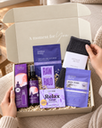 A curated self-care gift box opened on a cozy sofa, containing purple and lavender skincare items, a dark gray knit sock, and a white card. Hands are visible holding the box, with a warm home interior in the background. Relaxing self-care gift box with wool socks, sleep bath soak, self-heating eye mask, Relax soap, pillow spray and Raw Halo chocolate.