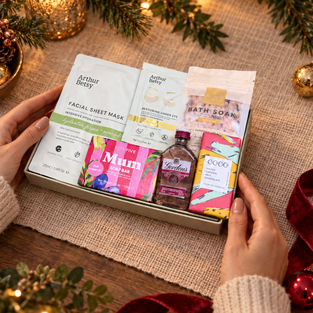 A Christmas-themed gift basket held by two hands, filled with skincare and self-care items including Arthur Beauty masks, a bath soak pouch, a bottle of lavender oil, and a bar of soap, surrounded by festive lights, pine branches, and a lit candle. Hallby letterbox gift for Mum with pink gin, salted caramel chocolate, bath soak and pampering face masks