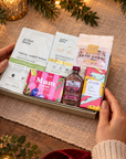 A Christmas-themed gift basket held by two hands, filled with skincare and self-care items including Arthur Beauty masks, a bath soak pouch, a bottle of lavender oil, and a bar of soap, surrounded by festive lights, pine branches, and a lit candle. Hallby letterbox gift for Mum with pink gin, salted caramel chocolate, bath soak and pampering face masks