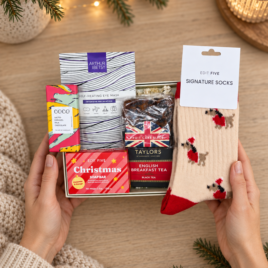 A gift set held by two hands on a wooden surface, featuring a box of Eye4 You eye wash, Dodo hand wash, Taylor's English Breakfast tea, a Christmas Sipsas box, and a pair of red and beige signature socks with a holiday tree design. The scene is decorated with pine branches, warm fairy lights, and a lit candle in the background. Hallby Christmas letterbox gift for her with festive Dachshund socks, tea, fruit cake, chocolate and self-care treats