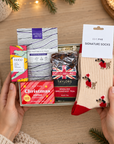 A gift set held by two hands on a wooden surface, featuring a box of Eye4 You eye wash, Dodo hand wash, Taylor's English Breakfast tea, a Christmas Sipsas box, and a pair of red and beige signature socks with a holiday tree design. The scene is decorated with pine branches, warm fairy lights, and a lit candle in the background. Hallby Christmas letterbox gift for her with festive Dachshund socks, tea, fruit cake, chocolate and self-care treats