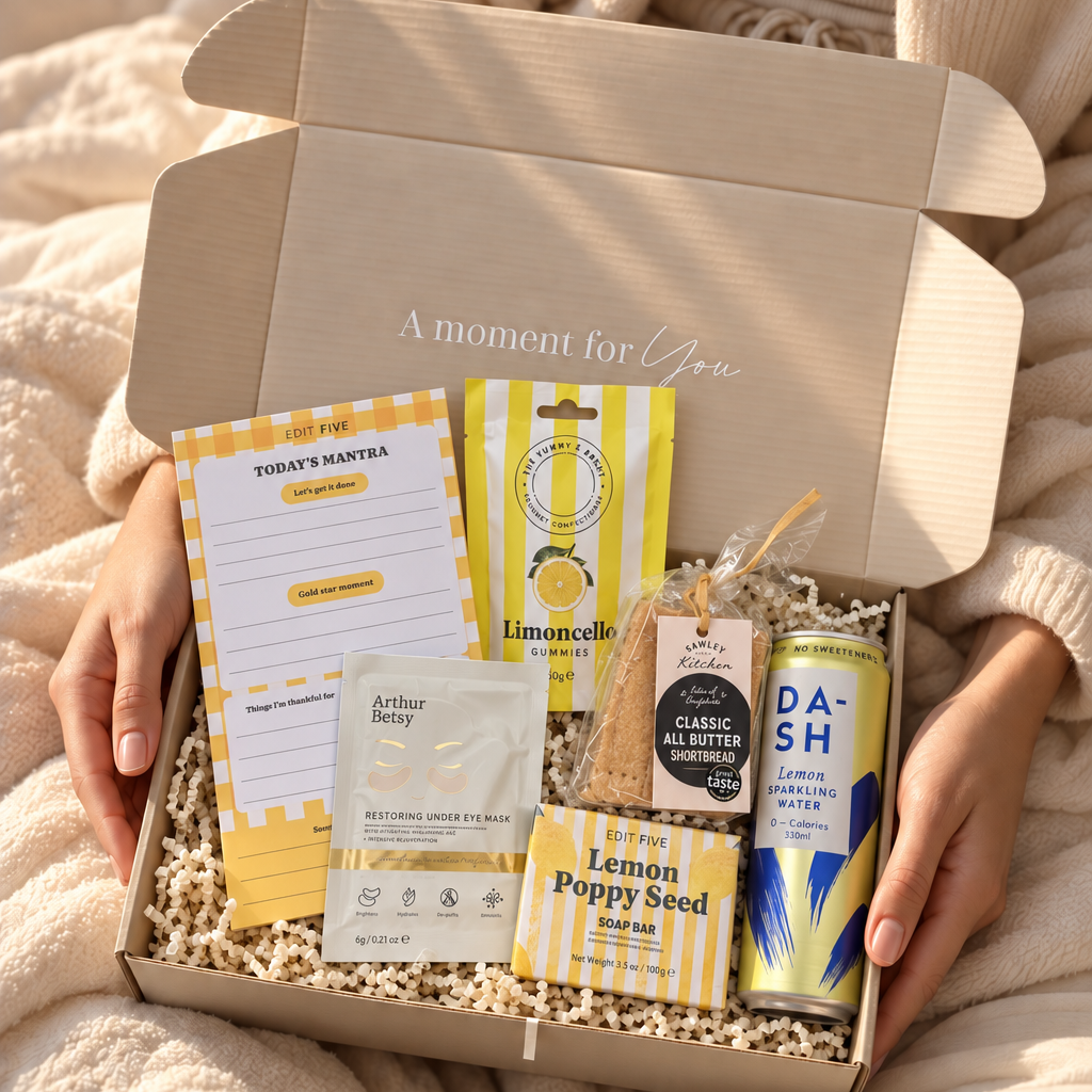 A person in a cream knit sweater holds an open gift box filled with wellness and snack items on a soft blanket. The box contains a yellow-and-white notepad, a lemon-themed card, a small packaged item, a tea bag, a can of DA-Sh lemonade, and a lemon poppy seed packet. Hallby cheer up gift hamper for her with lemon treats, positive notepad, soap and gold under-eye mask
