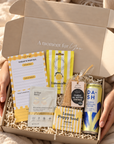 A person in a cream knit sweater holds an open gift box filled with wellness and snack items on a soft blanket. The box contains a yellow-and-white notepad, a lemon-themed card, a small packaged item, a tea bag, a can of DA-Sh lemonade, and a lemon poppy seed packet. Hallby cheer up gift hamper for her with lemon treats, positive notepad, soap and gold under-eye mask
