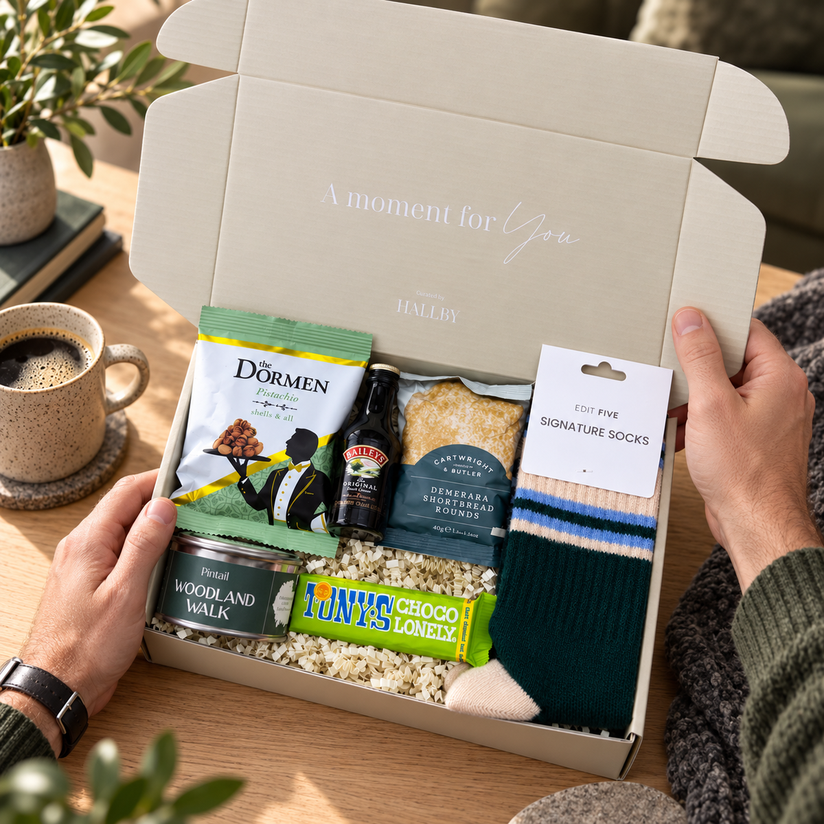 A gift box set held open by two hands on a wooden table, containing snacks and a pair of socks. Visible items include a bag of Dörmen chips, a bottle of Bailey's, a pack of Domestics bread, a 10ney chocolate bar, and a pair of dark green and cream signature socks. Christmas wellbeing gift for him with Green, Peach and Blue socks, chocolate, pistachios, shortbread and Baileys