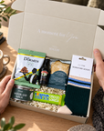A gift box set held open by two hands on a wooden table, containing snacks and a pair of socks. Visible items include a bag of Dörmen chips, a bottle of Bailey's, a pack of Domestics bread, a 10ney chocolate bar, and a pair of dark green and cream signature socks. Christmas wellbeing gift for him with Green, Peach and Blue socks, chocolate, pistachios, shortbread and Baileys