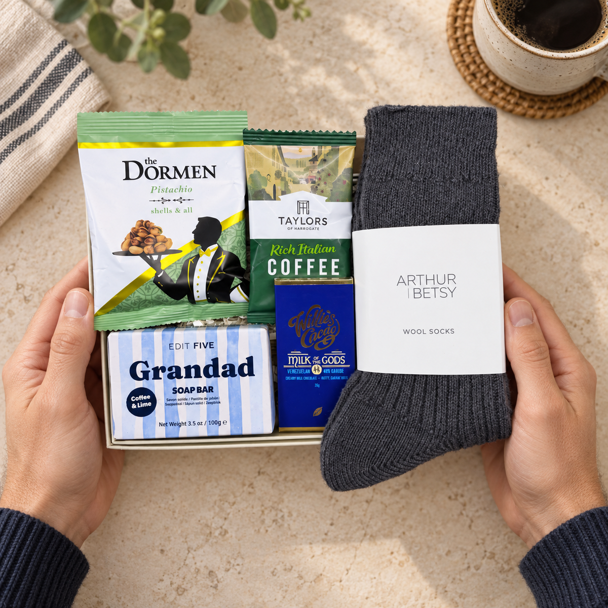 A gift box held by two hands contains curated items: a pair of dark charcoal wool socks with a white label reading Arthur Betsy, a bag of The Dormen coffee, a box of Taylor's Rich Indian Coffee, a blue box of Little &amp; Co's, and a striped soap bar labeled Grandad. The scene is styled on a beige stone surface with a mug and plant in the background. Letterbox gift for Grandad containing Taylors coffee, The Dormen pistachios and Willie's Milk of the Gods chocolate