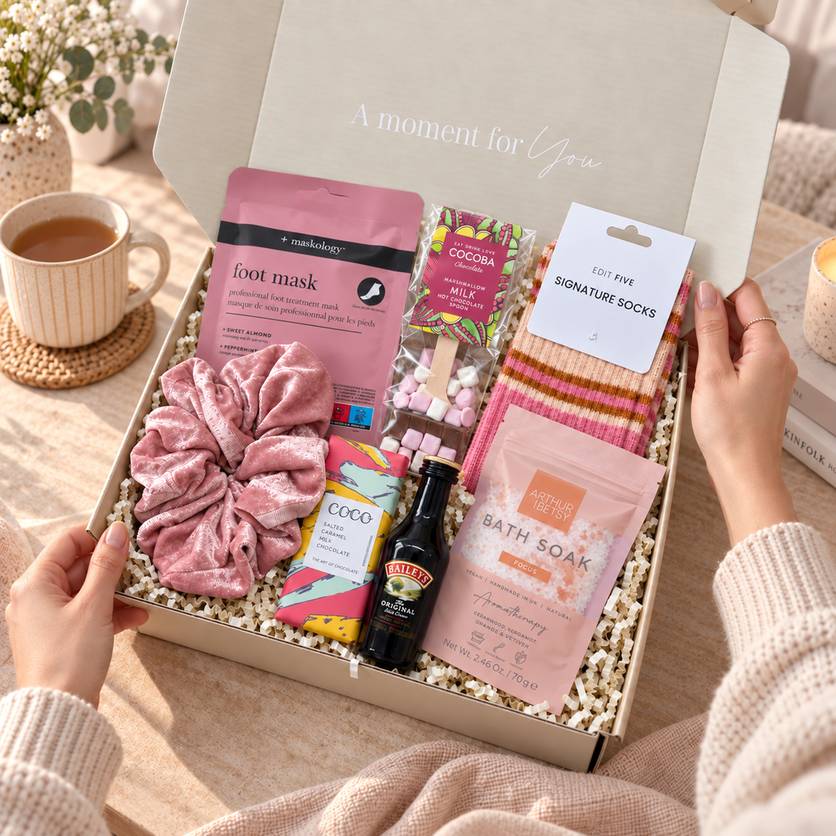 A curated gift box set opened by two hands, featuring a pink velvet scrunchie, a pink foot mask packet, a pink bath soak packet, striped socks, a chocolate bar, a small bottle, and a card labeled signature socks, arranged in white crinkle paper inside a light box with the text 'A moment for You'. Hallby Burr Box Christmas gift for her with colourful socks, hot chocolate, bath soak, foot mask and salted caramel chocolate