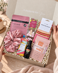 A curated gift box set opened by two hands, featuring a pink velvet scrunchie, a pink foot mask packet, a pink bath soak packet, striped socks, a chocolate bar, a small bottle, and a card labeled signature socks, arranged in white crinkle paper inside a light box with the text 'A moment for You'. Hallby Burr Box Christmas gift for her with colourful socks, hot chocolate, bath soak, foot mask and salted caramel chocolate