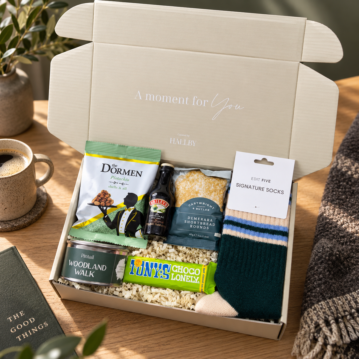 A curated gift box set from Ha'elys on a wooden table, featuring a cream-colored box with the text 'A moment for You' on the inside lid, filled with gourmet snacks, a can of wild woodland walk, a bottle of Baileys, a bag of Dormen, a bar of chocolate, and a pair of signature socks. Curated Christmas gift box for Dad featuring colourful socks, Pintail candle tin and indulgent food and drink treats
