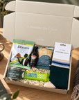 A curated gift box set from Ha'elys on a wooden table, featuring a cream-colored box with the text 'A moment for You' on the inside lid, filled with gourmet snacks, a can of wild woodland walk, a bottle of Baileys, a bag of Dormen, a bar of chocolate, and a pair of signature socks. Curated Christmas gift box for Dad featuring colourful socks, Pintail candle tin and indulgent food and drink treats
