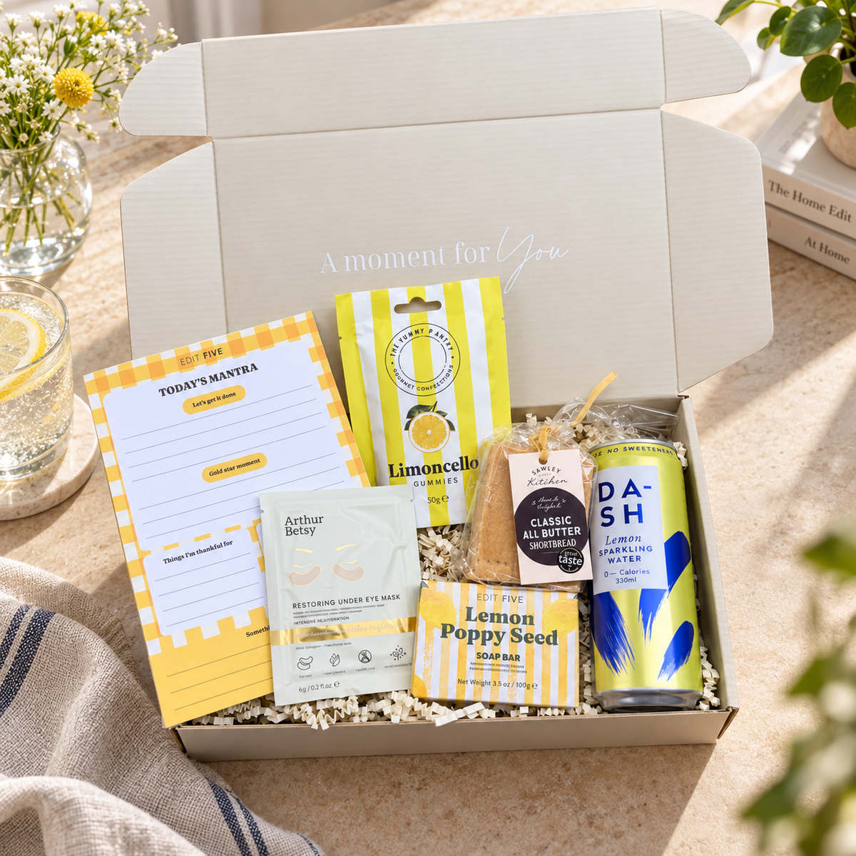 A curated gift box set displayed in an open white cardboard box with the lid propped open. The set includes a yellow-and-white notepad, a bag of Limoncello lemon balm, a packet of Arthur Betty retinol eye gel, a bag of lemon poppy seed, a small tag, and a can of Dash lemon sparkling water. Get well soon gift for her with Today’s Mantra notepad, DASH lemon water and uplifting self-care treats