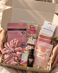 A curated gift box held by hands in a cozy setting, filled with pink and multicolor self-care items including a pink velvet scrunchie, foot mask packet, and other wellness accessories. Cosy hamper for women with Edit Five Pink, Peach and Orange socks, self-care products and festive treats