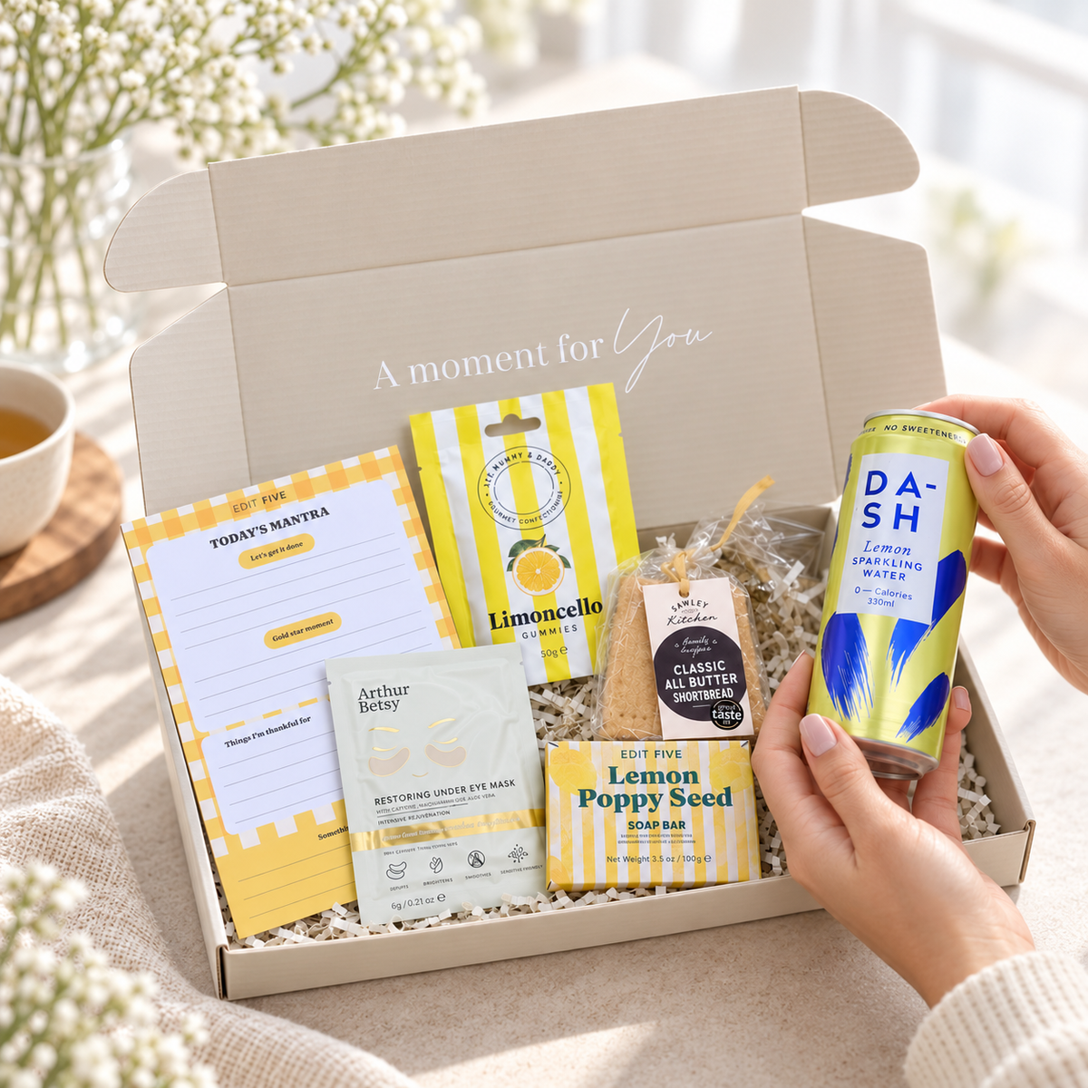 A curated gift box set displayed in an open beige box with a hand holding a can of Dash sparkling water. The box contains a yellow-and-white notepad, a lemon meringue cookie, an Arthur Bailey eye mask, and a lemon poppy seed bar, set on a beige fabric surface with soft floral background decor. Colourful gift hamper containing limoncello gummies, all-butter shortbread, lemon soap and gold under-eye mask