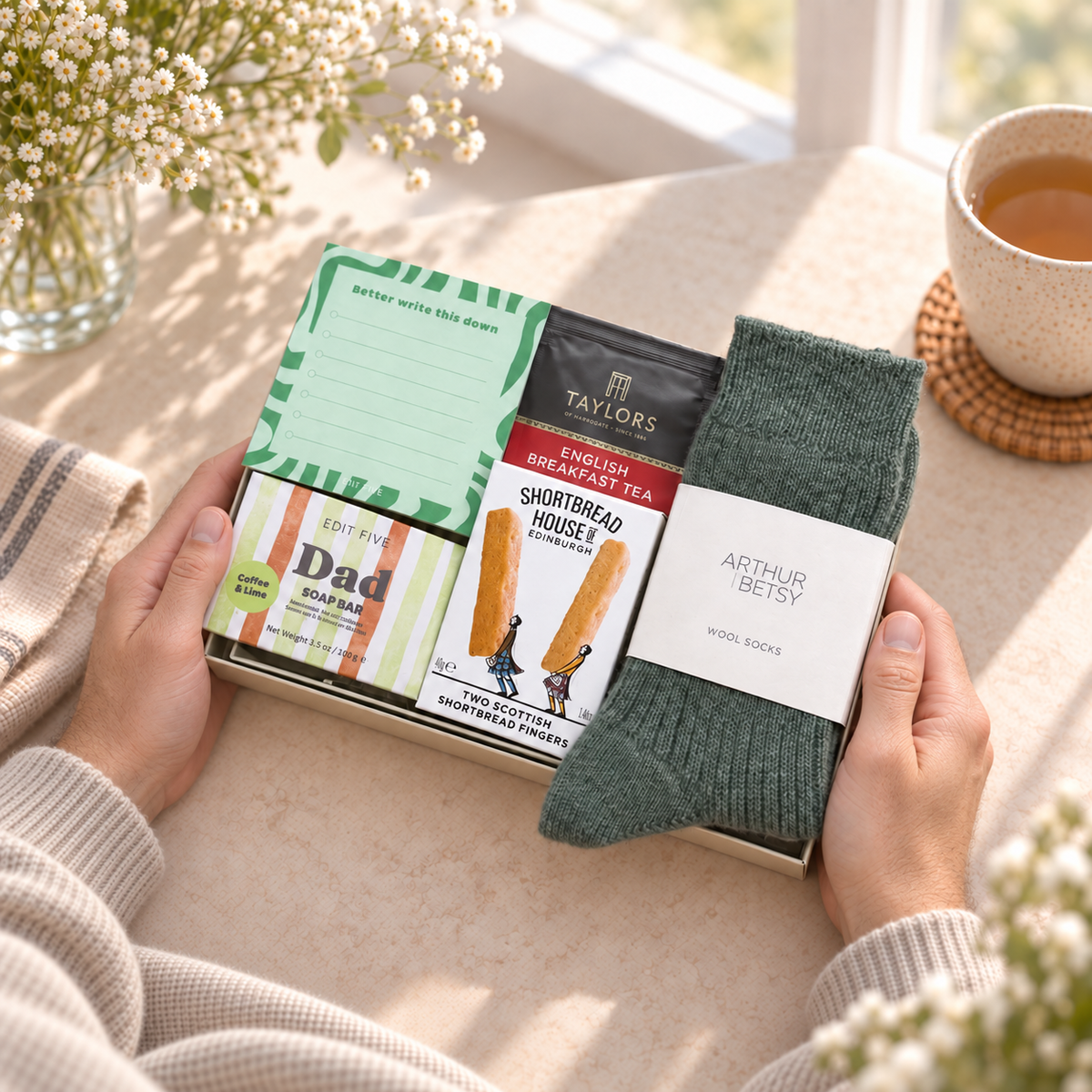 A gift box held by two hands on a sunlit table, containing a green notepad, a bag of Taylors English Breakfast Tea, a bar of soap, and a pair of dark green knit socks with a white Arthur Betsy label. A cup of tea and flowers are visible in the background. 
Hallby letterbox gift for Dad with Twilight wool socks, Dad soap, shortbread, English Breakfast tea and a Better Write This Down memo pad