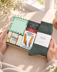 A gift box held by two hands on a sunlit table, containing a green notepad, a bag of Taylors English Breakfast Tea, a bar of soap, and a pair of dark green knit socks with a white Arthur Betsy label. A cup of tea and flowers are visible in the background. 
Hallby letterbox gift for Dad with Twilight wool socks, Dad soap, shortbread, English Breakfast tea and a Better Write This Down memo pad