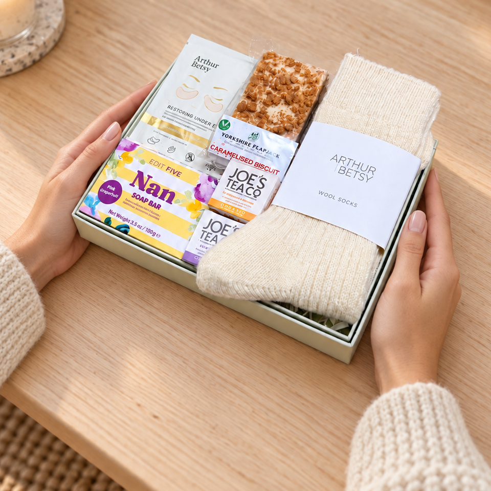 A gift box held by two hands on a wooden table, containing a cream knit sock, a bar of soap, a small packaged item, and a wrapped snack. The scene is styled with warm holiday lights, candles, and books in the background. Hallby letterbox gift for Nan with wool socks, Nan soap bar, Gold under-eye patches, Joe’s Tea Co. tea and caramelised biscuit flapjack