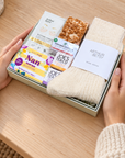 A gift box held by two hands on a wooden table, containing a cream knit sock, a bar of soap, a small packaged item, and a wrapped snack. The scene is styled with warm holiday lights, candles, and books in the background. Hallby letterbox gift for Nan with wool socks, Nan soap bar, Gold under-eye patches, Joe’s Tea Co. tea and caramelised biscuit flapjack