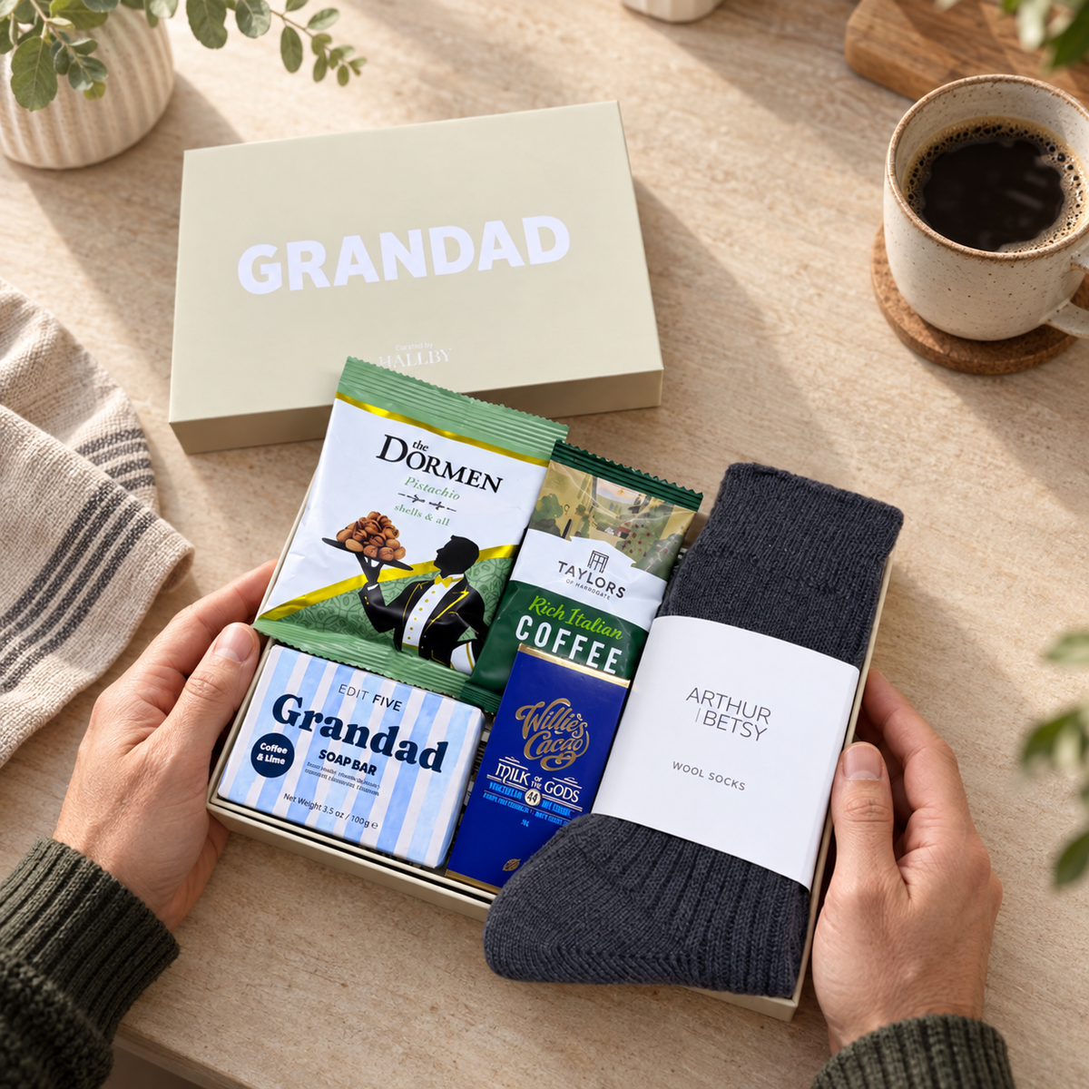 A gift box opened by two hands on a beige tabletop, containing a dark gray knit sock labeled Arthur Betsy, a boxed soap bar labeled Grandad, and two coffee bags. A matching beige gift box lid with the word GRANDAD sits behind it, with a cup of coffee and plants in the background. Thoughtful Grandad gift hamper with Arthur Betsy Twilight socks and Edit Five Grandad soap bar