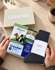 A gift box opened by two hands on a beige tabletop, containing a dark gray knit sock labeled Arthur Betsy, a boxed soap bar labeled Grandad, and two coffee bags. A matching beige gift box lid with the word GRANDAD sits behind it, with a cup of coffee and plants in the background. Thoughtful Grandad gift hamper with Arthur Betsy Twilight socks and Edit Five Grandad soap bar